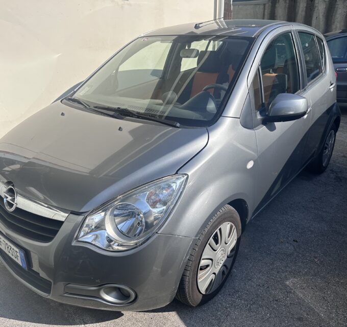 Front three-quarter view of a silver car parked beside a light wall, with another car in the background; license plate visible on the front bumper.