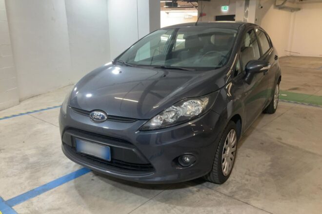Gray Ford sedan parked in an indoor parking garage, front-left view showing headlights and grille.