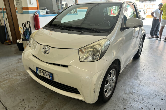 Front-angle view of a white Toyota parked inside a service bay, with a red fire extinguisher on the wall and people standing near the entrance in the background.