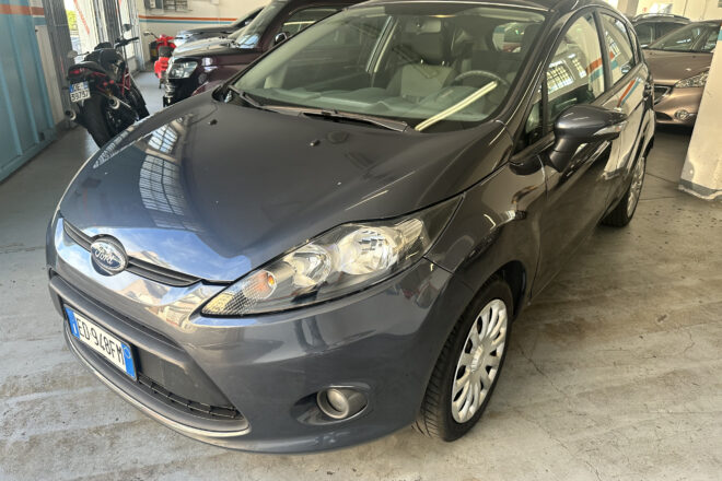Front-left view of a dark gray Ford sedan parked in a bright indoor garage with other cars and a blue-striped wall in the background.