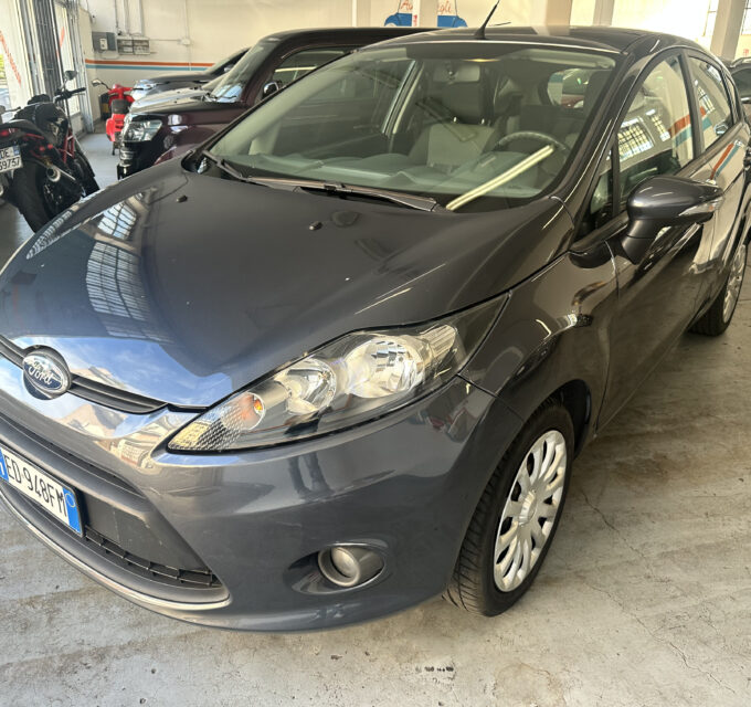 Front-left view of a dark gray Ford sedan parked in a bright indoor garage with other cars and a blue-striped wall in the background.