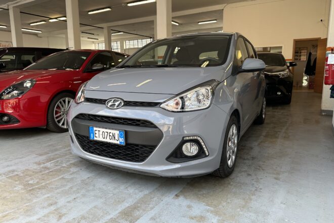 Grey Hyundai hatch in a bright indoor showroom or garage, with a red car parked to its left and other vehicles in background.