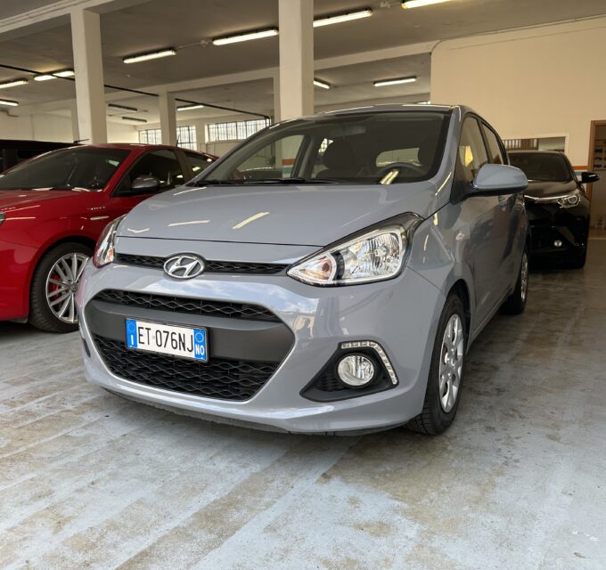 Grey Hyundai hatch in a bright indoor showroom or garage, with a red car parked to its left and other vehicles in background.