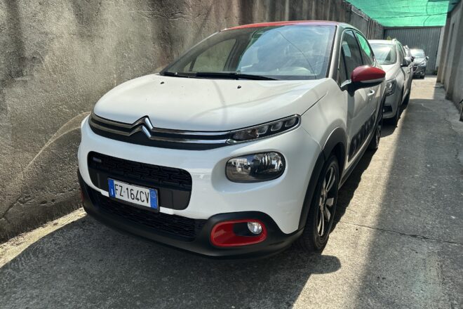 White Citroën SUV parked in a narrow alley beside a weathered wall, with a green shade sail overhead.