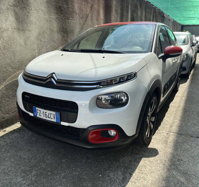 White Citroën SUV parked in a narrow alley beside a weathered wall, with a green shade sail overhead.