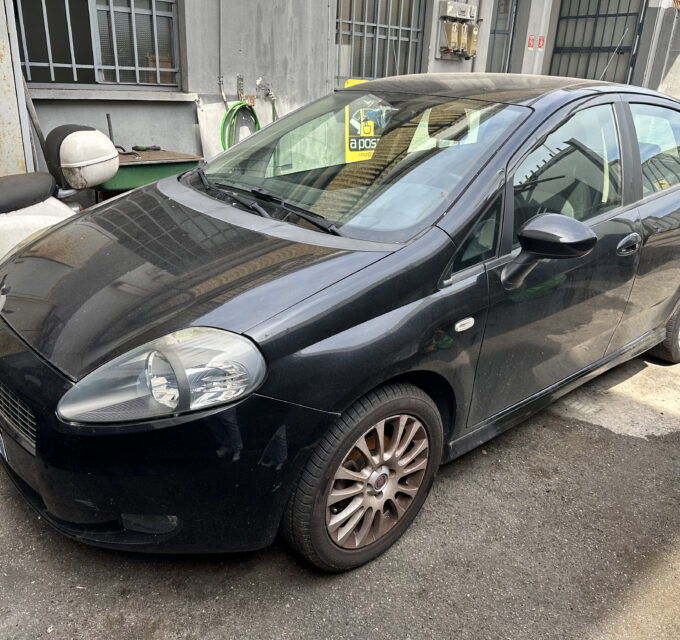 Dark-colored compact car (front-left) parked in an urban alley beside a weathered metal door and garden hose reels on the wall behind it.