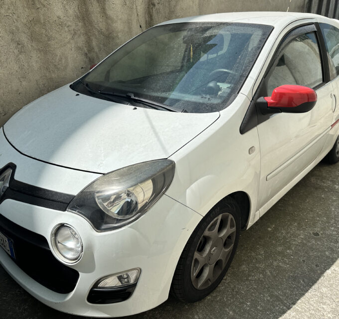 White compact car parked beside a rough concrete wall; red side mirror stands out on the driver's side.