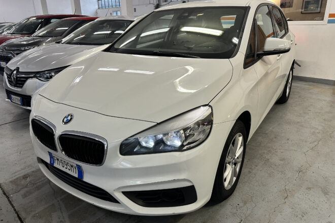 Front-left view of a white BMW SUV in a dealership showroom, with other cars lined up behind it.