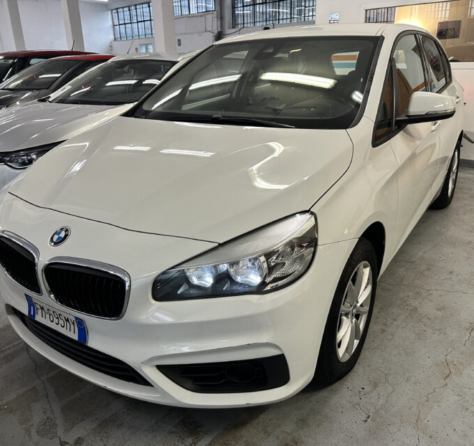 Front-left view of a white BMW SUV in a dealership showroom, with other cars lined up behind it.
