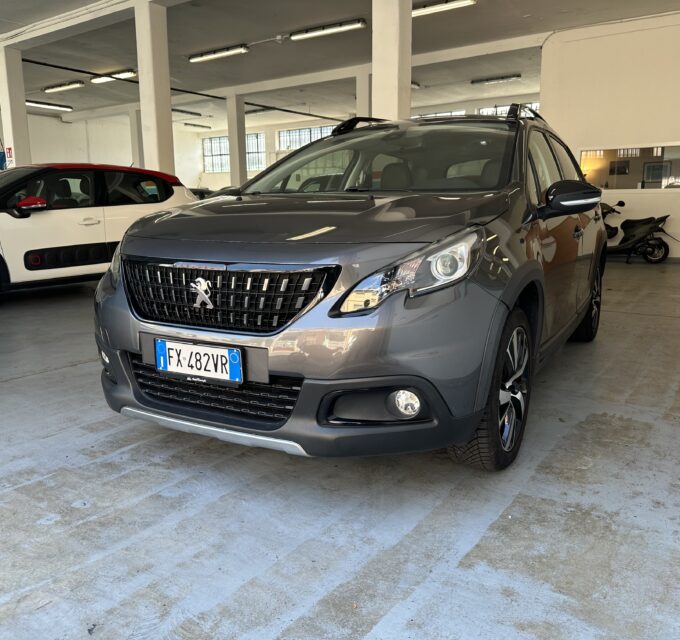 Indoor car showroom with a gray Peugeot SUV in the foreground and a white and red compact car to the left; showroom sign 'AutoNavigi' in the background.