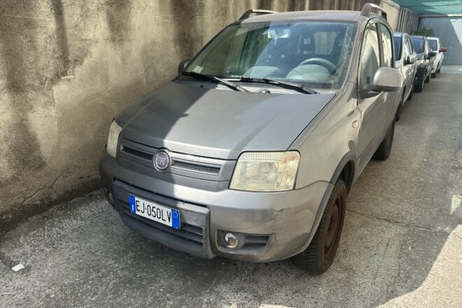Silver Fiat van in the foreground parked along a concrete wall, with a line of cars behind under a green canopy.