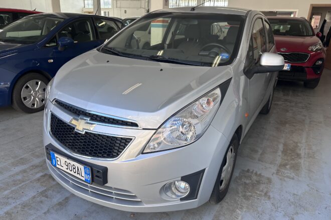 Silver Chevrolet SUV in the foreground inside an indoor car showroom/garage, with a blue car to the left and a red Kia in the background.