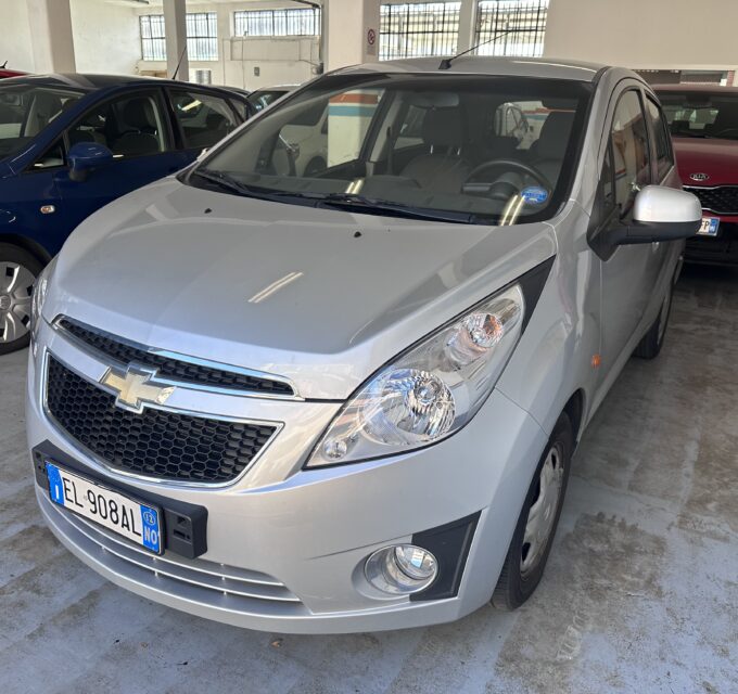 Silver Chevrolet SUV in the foreground inside an indoor car showroom/garage, with a blue car to the left and a red Kia in the background.