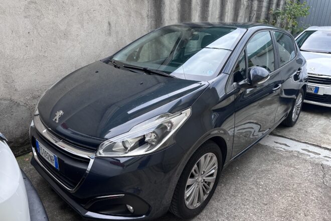 Front-left view of a dark gray Peugeot hatchback parked beside a rough concrete wall in a parking area.