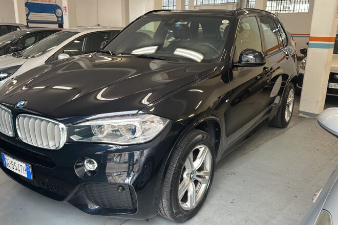 Black BMW SUV parked in an indoor garage among other cars, front grille and headlights visible, glossy finish reflects overhead lights