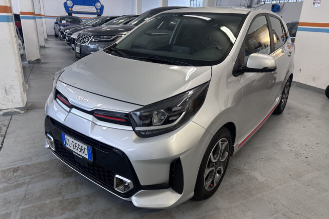 Front three-quarter view of a silver Kia minivan in a showroom with other cars lined up in the background; wall mural says AutoNavigli with a blue car drawing.