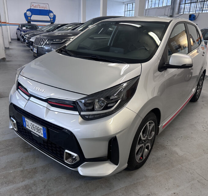 Front three-quarter view of a silver Kia minivan in a showroom with other cars lined up in the background; wall mural says AutoNavigli with a blue car drawing.