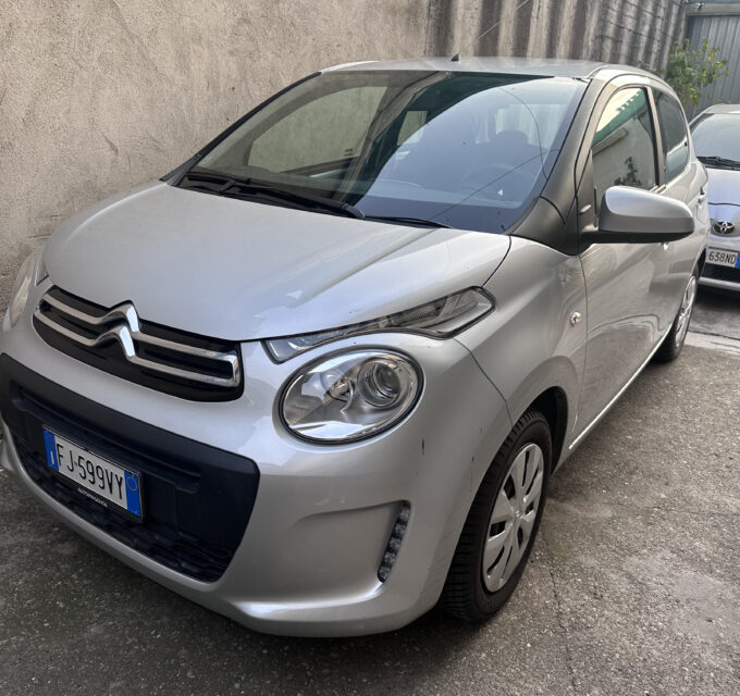 Front left of a silver Citroën car parked beside a beige wall, with another white car in the background.