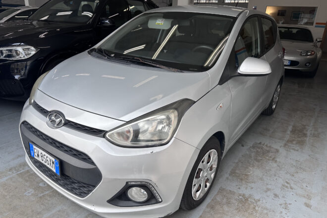 Front three-quarter view of a silver Hyundai in an indoor parking area with other cars nearby and polished concrete floor. Front bumper and grille visible, license plate partially shown.