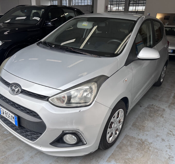 Front three-quarter view of a silver Hyundai in an indoor parking area with other cars nearby and polished concrete floor. Front bumper and grille visible, license plate partially shown.