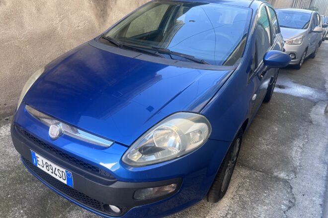 Blue Fiat car parked on a street with a tan wall in the background, other cars behind it partially visible.