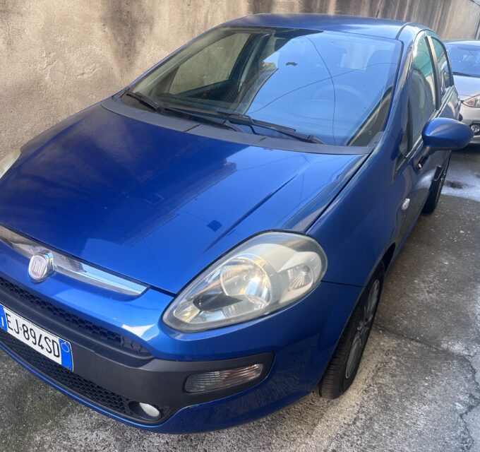 Blue Fiat car parked on a street with a tan wall in the background, other cars behind it partially visible.