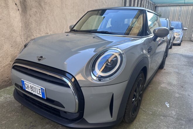 Front-left view of a gray Mini Cooper parked in a narrow lot, with a beige wall on the left and other cars in line behind it.