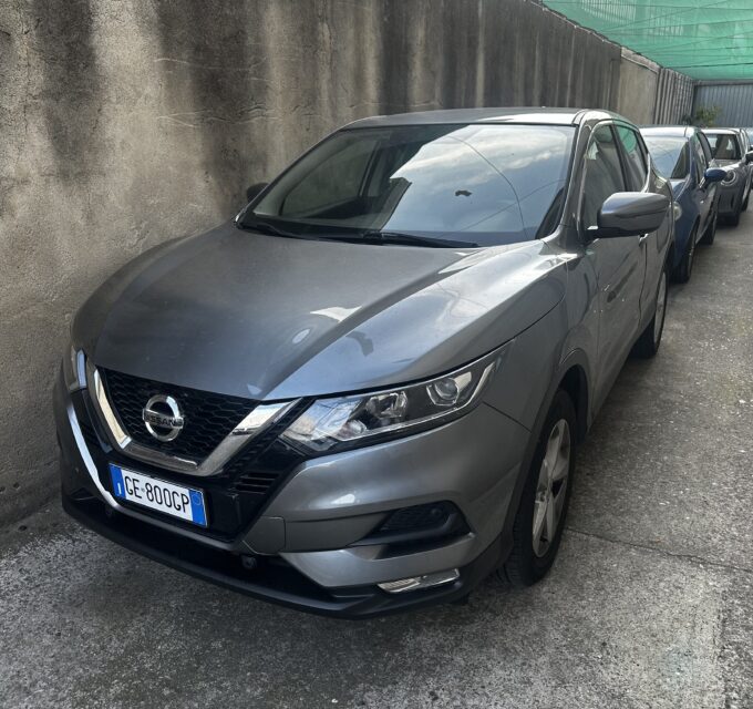 Line of parked cars along a narrow alley with a tall concrete wall on the left, gray SUV in foreground and more cars behind, green netting overhead.