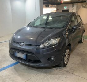Gray Ford sedan parked in an indoor parking garage, front-left view showing headlights and grille.