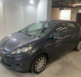 Gray Ford Fiesta hatchback parked in an indoor parking garage with concrete columns and overhead lights on a marked parking spot with blue lines in the floor journey area.