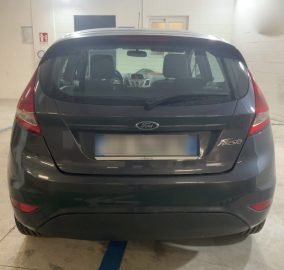 Rear view of a dark gray Ford Fiesta hatchback parked in a well-lit indoor garage, license plate blurred for privacy, taillights visible.