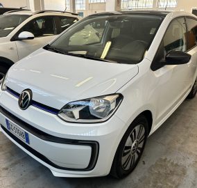 White Volkswagen hatchback parked indoors among other cars in a showroom, front angled view showing the grille and headlights.