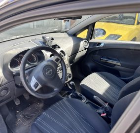 Interior of a car showing the driver’s seat, steering wheel, dashboard, and gearstick; a yellow car is visible through the passenger window outside.