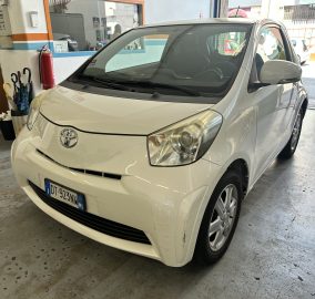 Front-angle view of a white Toyota parked inside a service bay, with a red fire extinguisher on the wall and people standing near the entrance in the background.