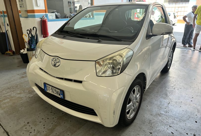 Front-angle view of a white Toyota parked inside a service bay, with a red fire extinguisher on the wall and people standing near the entrance in the background.