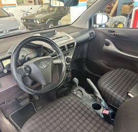 Interior of a car showing steering wheel, dashboard, center console, and patterned cloth seats; view through the windshield to a showroom with other vehicles outside