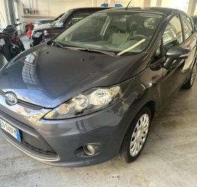 Front-left view of a dark gray Ford sedan parked in a bright indoor garage with other cars and a blue-striped wall in the background.