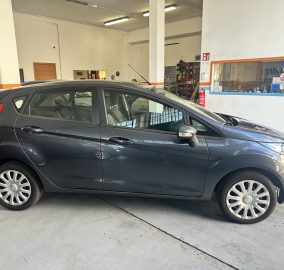 Dark gray hatchback parked inside a garage showroom with white walls and orange-and-blue trim, other cars visible in background.