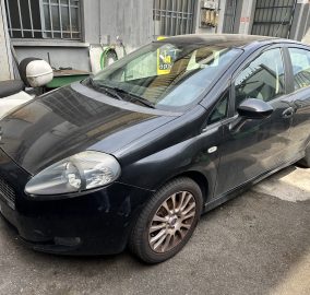 Dark-colored compact car (front-left) parked in an urban alley beside a weathered metal door and garden hose reels on the wall behind it.