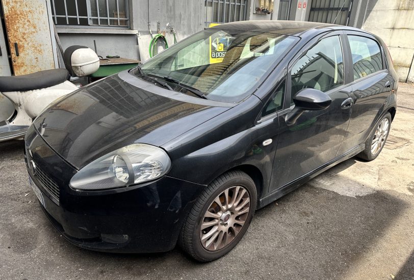 Dark-colored compact car (front-left) parked in an urban alley beside a weathered metal door and garden hose reels on the wall behind it.