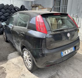 Black Fiat hatchback parked beside a tall stack of tires in an industrial yard, rear view visible.