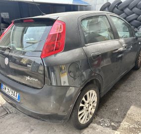 Dusty black Fiat Punto parked next to a tall pile of tires in an outdoor lot, rear and side view visible.