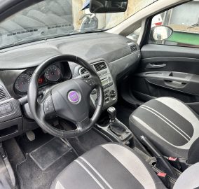 Interior of a Fiat car with steering wheel and dashboard; dusty surfaces, gear shift, and front seats visible; outside shows a green dumpster/metal container.