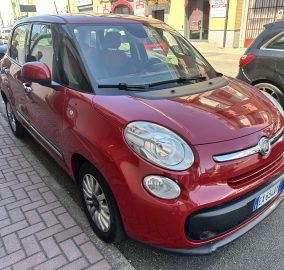 Red Fiat minivan parked on a city street, front-right quarter view with storefronts in the background, reflections on the glossy body.