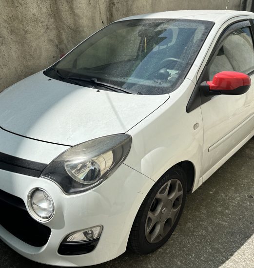 White compact car parked beside a rough concrete wall; red side mirror stands out on the driver's side.