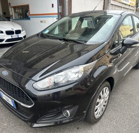 Front-left perspective of a dusty black Ford car parked on a city street, with a white BMW inside a garage to the left and other cars in the background