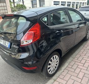 Black hatchback parked on a city street, showing the rear and taillight with a license plate visible (European plate). It’s beside a sidewalk with buildings in the background.