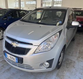 Silver Chevrolet SUV in the foreground inside an indoor car showroom/garage, with a blue car to the left and a red Kia in the background.