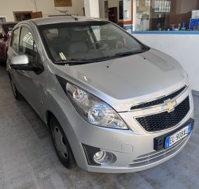 Front three-quarter view of a silver Chevrolet SUV parked indoors, with another red car behind it and a blue-framed window to the right.