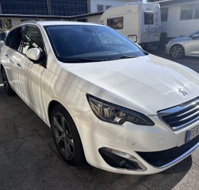 White Peugeot minivan parked in a car lot with a camper trailer and other vehicles in the background, taken from a front-side angle.